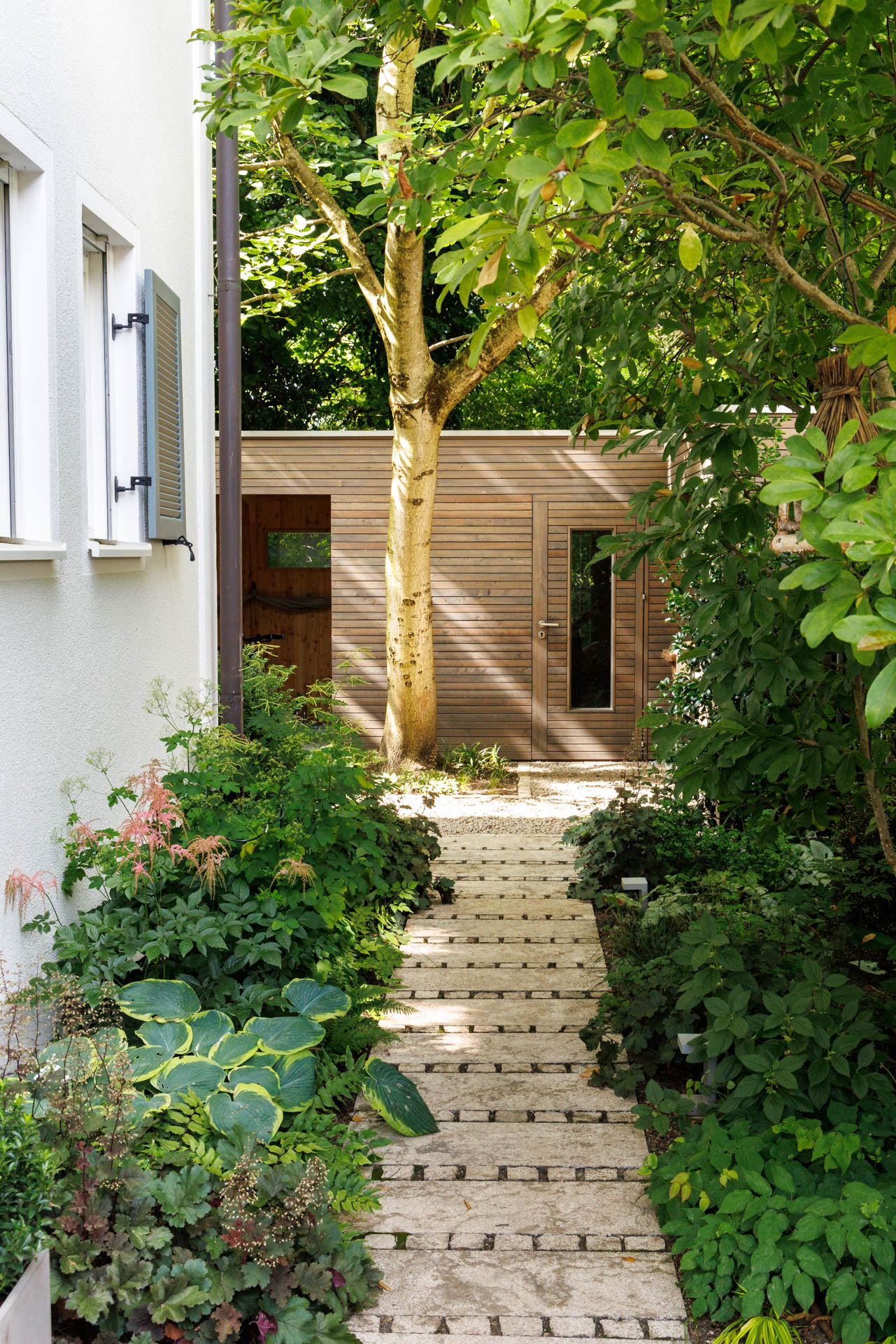 Der Gartenweg führt am Haus vorbei in den rückwärtigen Garten. Ganz hinten steht das Gartenhaus aus Europäischer Lärche mit Rhombus-Fassade. Schattenstauden säumen das Bild.
