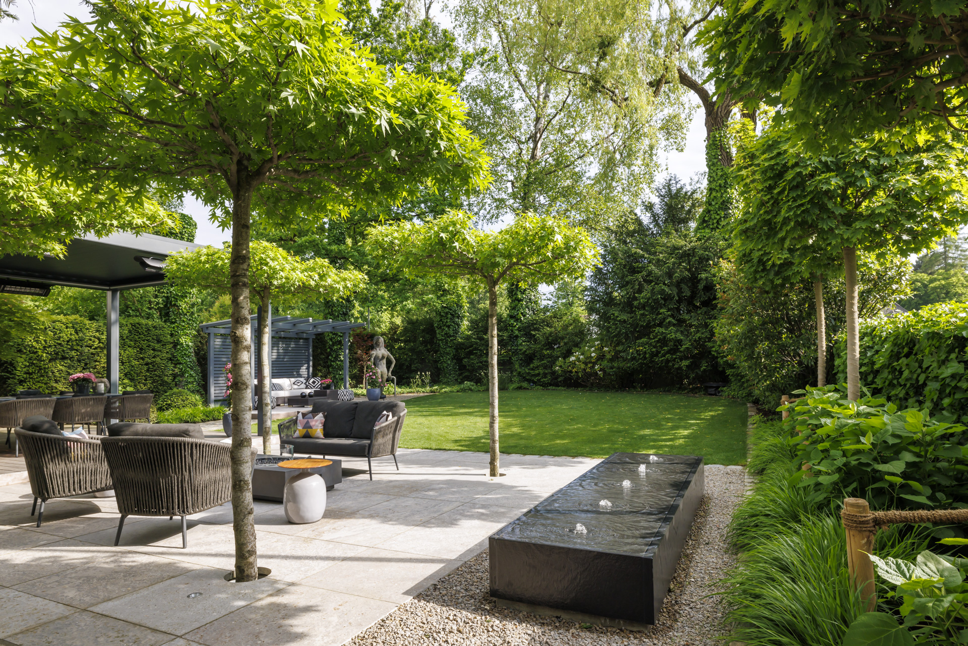 Blick über den Wassertisch, mit Oase-Pumpen, Seliger Quellstar Beleuchtung, über die Terrasse mit den dachförmig geschnittenen Bäumen.