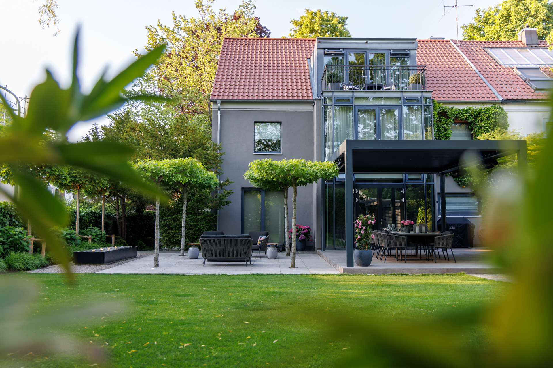 Blick zurück aus dem Garten auf das Wohnhaus. Die zwei Terrassen: Lounge Muschelkalk und Hauptterrasse Holzdeck aus Ipé liegen davor. Die Rasenfläche schließt sich an.